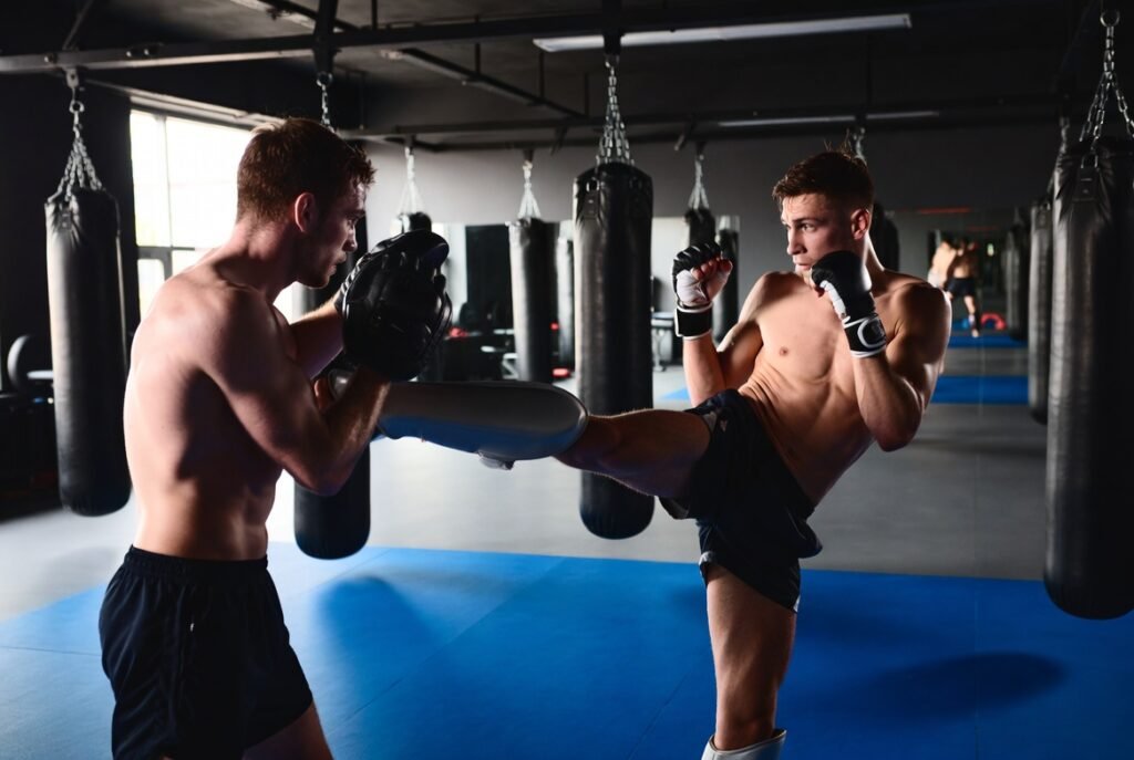 Need Shin Guards for MMA and Kickboxing 7 Powerful Reasons 1 Fighter wearing shin guards performing kickboxing leg kick drill during MMA training in a gym with a partner holding pads, showing proper protective gear usage and controlled sparring technique.